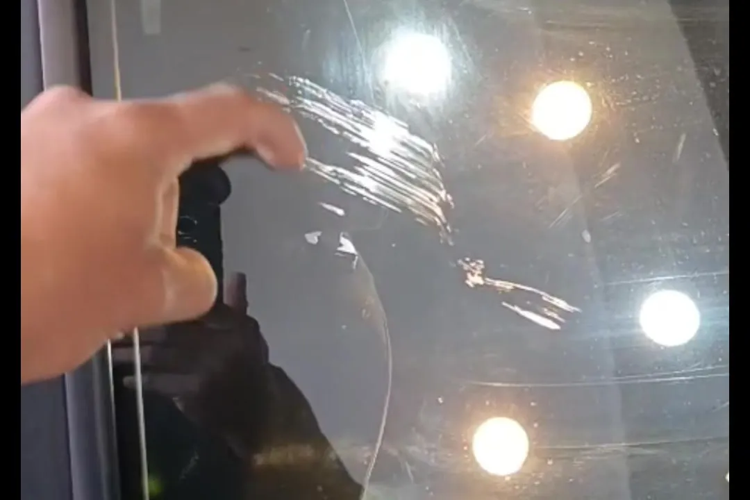 Vidro automotivo com riscos profundos, revelados pela luz da oficina, com uma mao apontando o dano