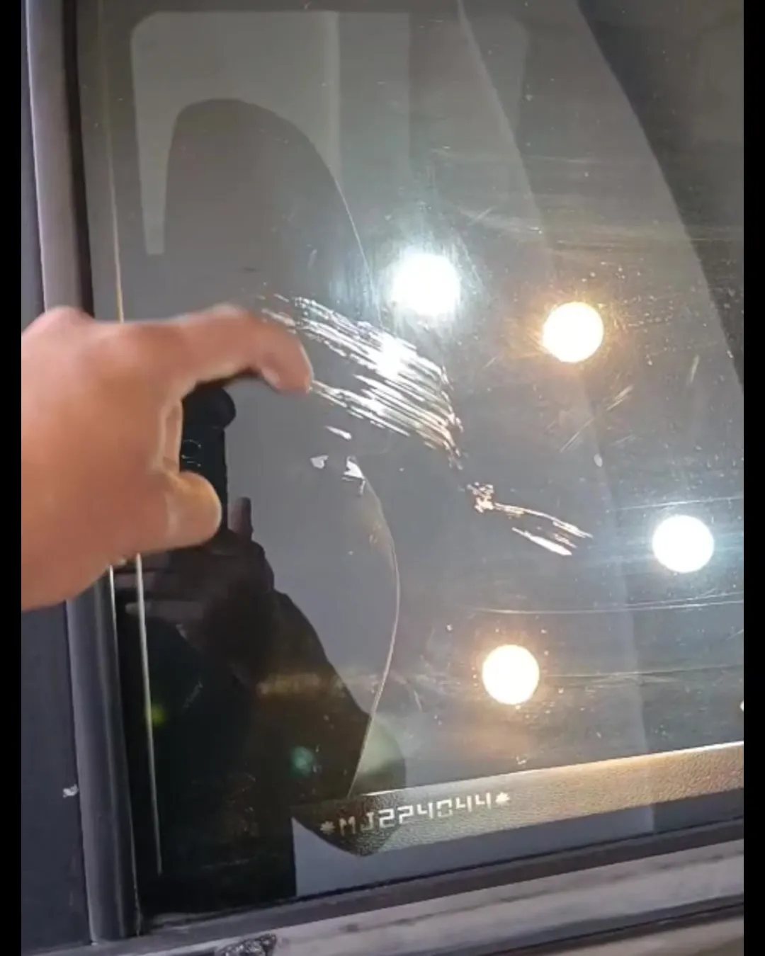 Vidro automotivo com riscos profundos, revelados pela luz da oficina, com uma mao apontando o dano
